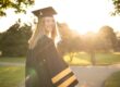 Graduate in cap and gown smiling outdoors with sunlight and trees in the background on a sunny day.