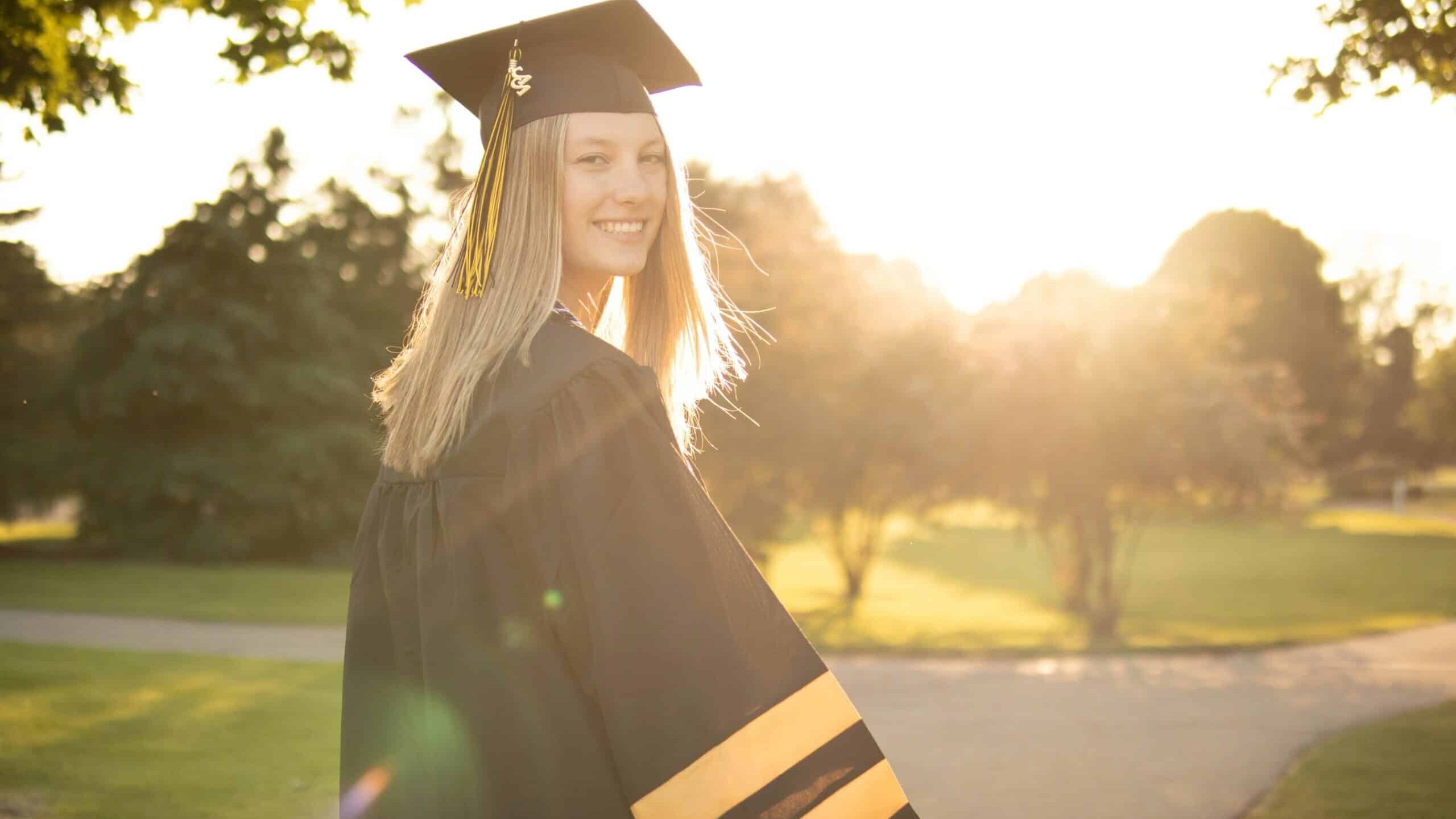 High School Grad Graduate in cap and gown smiling outdoors with sunlight and trees in the background on a sunny day.