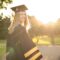 Graduate in cap and gown smiling outdoors with sunlight and trees in the background on a sunny day.