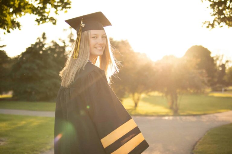 Graduate in cap and gown smiling outdoors with sunlight and trees in the background on a sunny day.
