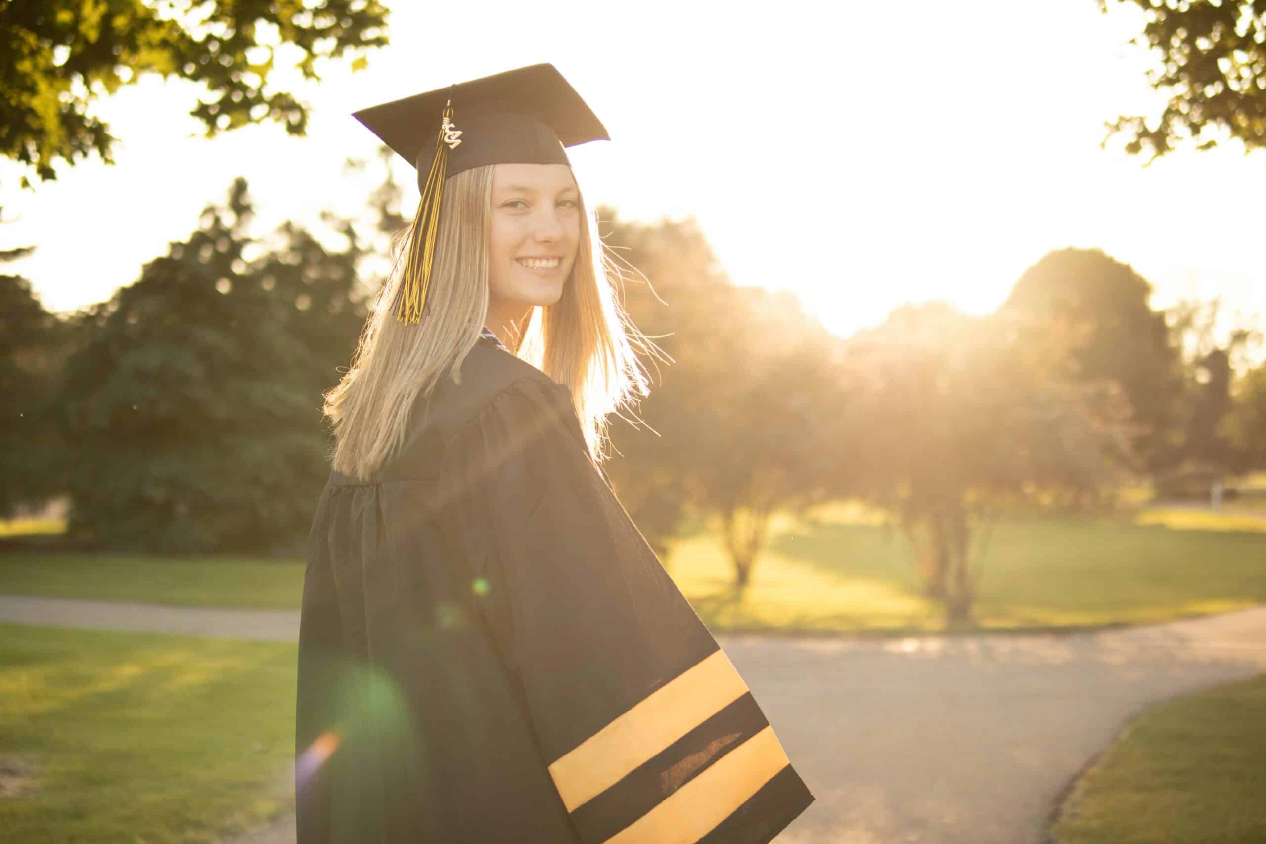Graduate in cap and gown smiling outdoors with sunlight and trees in the background on a sunny day.